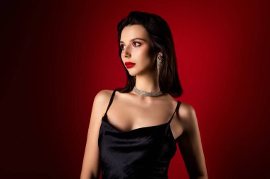 Şık siyah elbiseli, göz kamaştırıcı mücevherleri olan, esmer saçlı, büyüleyici kırmızı dudaklı, çarpıcı kırmızı arkaplana karşı zarif bir duruşu olan çekici bir genç kadın..