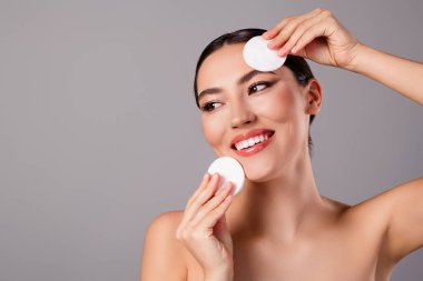 Kusursuz derisi olan, gülümseyen ve cilt bakımı için pamuk tutan, gri arka planda güzelliğini ve sağlığını gösteren genç bir kadın..