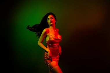 Şık, genç bir kadın model saten bir elbiseyle zarafet sergiliyor. Parlak neon ışıkla aydınlatılıyor. Güzelliğini vurguluyor..