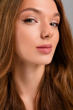 Dalgalı kahverengi saçlı, yumuşak makyajlı ve parlak tenli genç bir kadının yakın çekimi. Gri arkaplana karşı yeni, zarif bir görünüm sergiliyor..