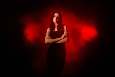 Şık bir genç kadın, kırmızı duman ve ışığın arasında, karanlık ve dramatik bir ortamda kendine güvenerek duruyor..