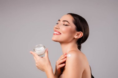Çekici esmer bir kadın cilt kremi sürmekten zevk alır. Parlak gülüşü ve kusursuz teni güzelliği ve özgüvenini ön plana çıkarır..