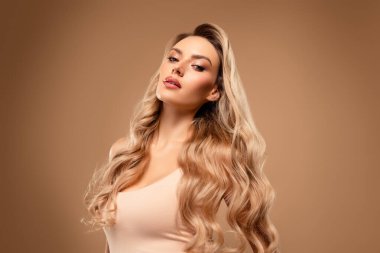 Güzel sarışın kadın çarpıcı dalgalı saç stili ve zarif güzelliğini sergiliyor, stüdyoda poz veriyor yumuşak bej bir arka planda şık giyiniyor, zarafet ve inceliği somutlaştırıyor..