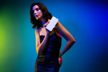 Güzel esmer kadın canlı mavi ve yeşil neon ışığın altında beyaz akıllı telefonuyla poz veriyor, gençlik zarafeti sergiliyor ve modern bir fotoğrafçılık ortamında büyüleyici bir parti estetiği sergiliyor..