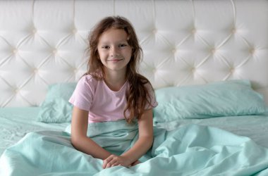 Küçük bir kız pijamalarıyla yatakta oturur ve kameraya gülümser..