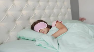 Küçük bir kız, yenilenmiş bir yatak odasında büyük bir yatakta uyku maskesiyle uyuyor. Sabahları uyanıp yeni bir güne başlama fikri. 4k görüntü