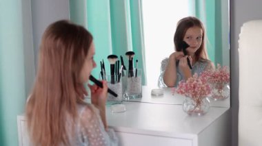 Makyaj yapıyormuş gibi yapan gülümseyen küçük bir kız makyaj fırçası tutuyor. Çocuk oyunu, güzellik figürleri, masumiyet, kendini ifade etme ve evde yaratıcılık. 4k görüntü