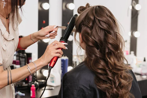 Mãos de um cabeleireiro com um ferro de ondulação fazendo um penteado para uma menina encaracolada — Fotografia de Stock