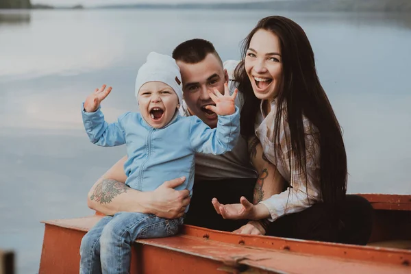 Молодая счастливая семья с сыном сидят и улыбаются в лодке у воды — стоковое фото