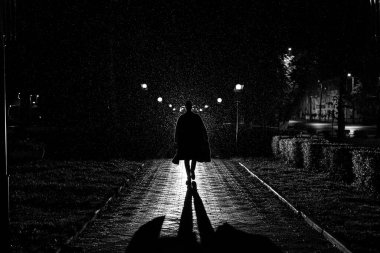 Gece vakti şehirde dolaşan şapkalı ve yağmurluklu bir adamın dramatik silueti.