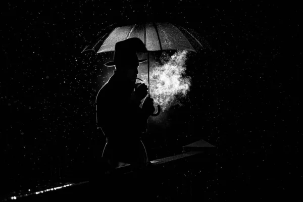 Silhueta de um homem de chapéu sob um guarda-chuva Fumar um cigarro à noite na chuva na cidade — Fotografia de Stock