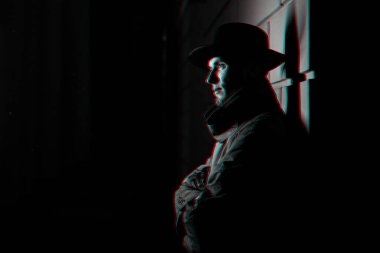 Gece sokakta yağmurluk giymiş, şapkalı bir adamın koyu renkli portresi.