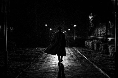 Gece sokağında yağmurda paltolu ve şapkalı bir erkek dedektifin karanlık silueti.