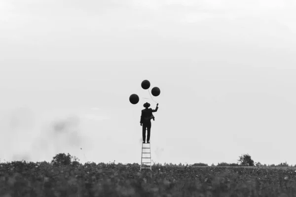 Foto surrealista de un hombre soltero en un traje en una escalera con globos en un campo —  Fotos de Stock