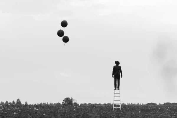 一个穿着西装站在楼梯上，拿着气球站在田野里的人的超现实的肖像 — 图库照片