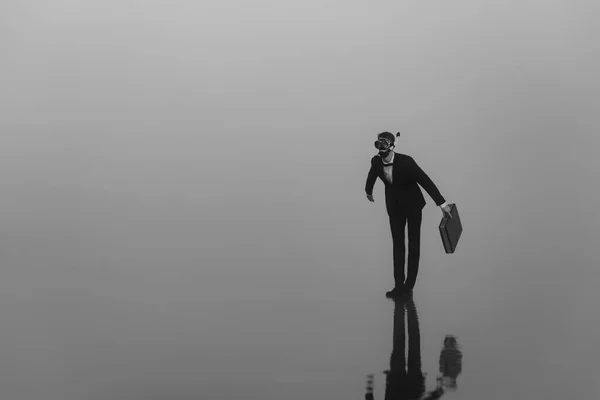 Surreales Porträt eines Mannes in Anzug und Maske mit Tauchschlauch und Aktentasche in den Händen, der auf dem Wasser steht — Stockfoto