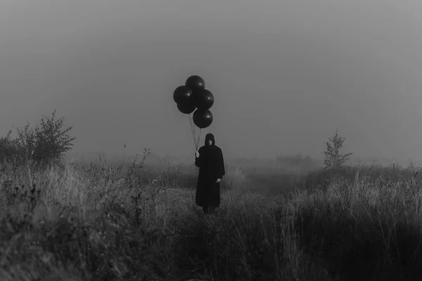 Spaventoso uomo scuro in un costume mostro con un cappuccio con palloncini in mano — Foto Stock