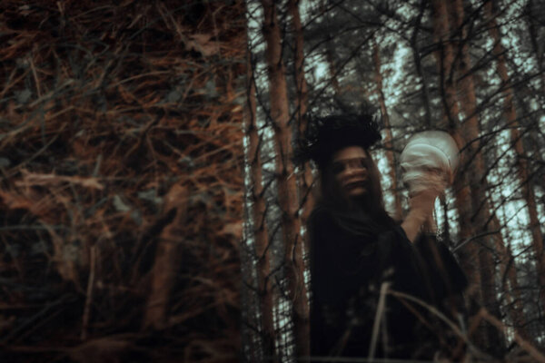 Blurry mystical reflection of a black witch with a skull in her hands performing a satanic ritual in a dark forest