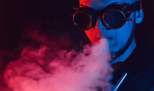Porträt eines Mannes mit futuristischer Brille raucht eine Wasserpfeife und pustet eine Rauchwolke in eine Shisha-Bar — Stockfoto