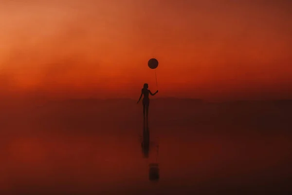 Surrealistyczna sylwetka dziewczyny z balonem w dłoni na wodzie o wschodzie słońca we mgle w lecie — Zdjęcie stockowe