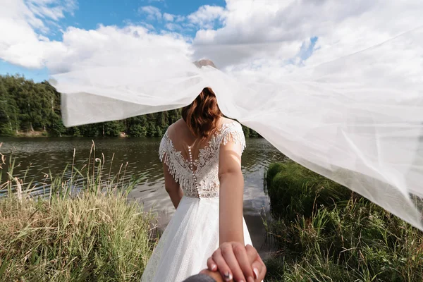 Esküvő kövessen koncepció. A menyasszony fogja a vőlegény kezét az esküvő napján a természetben a folyónál. — Stock Fotó