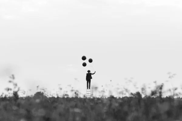 Foto surrealista de un hombre soltero en un traje en una escalera con globos en un campo —  Fotos de Stock