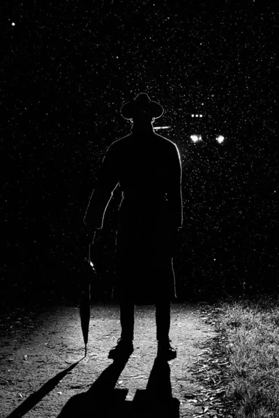 Silhouette sombre d'un homme en imperméable et chapeau sous un parapluie dans la rue sous la pluie — Photo