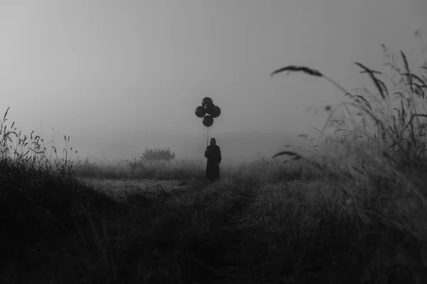 Hombre con un disfraz de un monstruo terrible en un cabo con una capucha se encuentra en la niebla en un campo —  Fotos de Stock