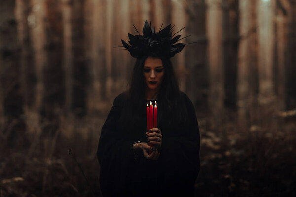 Black terrible witch holds candles in her hands in a dark forest
