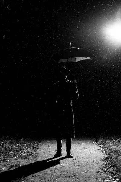 Silhueta escura de um homem em uma capa de chuva e chapéu sob um guarda-chuva na rua na chuva — Fotografia de Stock