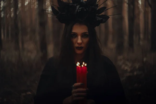 Strega in costume nero tiene le candele in una foresta scura — Foto Stock
