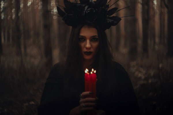 Witch in a black costume holds candles in a dark gloomy forest