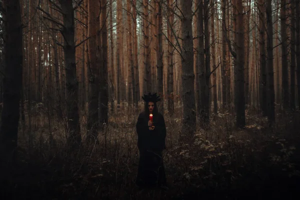 Bruja en un traje negro sostiene velas en un bosque oscuro — Foto de Stock