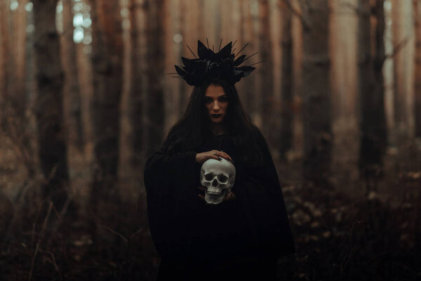 Black terrible witch with a skull in the hands of a dead man in the woods