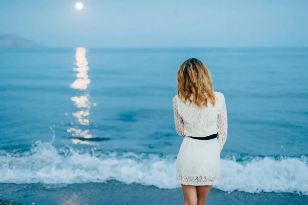 Chica en un vestido blanco está de pie con su espalda en la playa por el mar — Foto de Stock