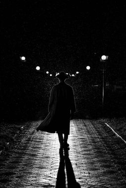 Gece vakti şehirde dolaşan şapkalı ve yağmurluklu bir adamın dramatik silueti.