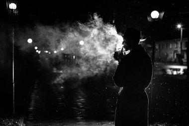 Gece sokağında yağmurda sigara içen şapkalı bir adamın karanlık silueti.
