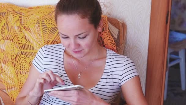 Casa, tecnologia e conceito de internet - mulher sorridente com smartphone sentado no sofá em casa — Vídeo de Stock