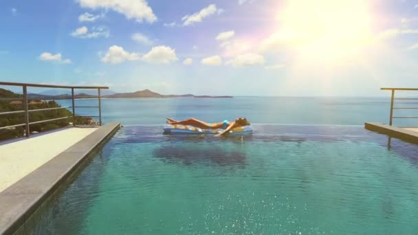 Style de vie en plein air portrait de jeune fille assez sexy nageant avec matelas lumineux néon à la piscine, portant un bikini et des lunettes de soleil, vue sur l'océan, se détendre et s'amuser en vacances . — Video