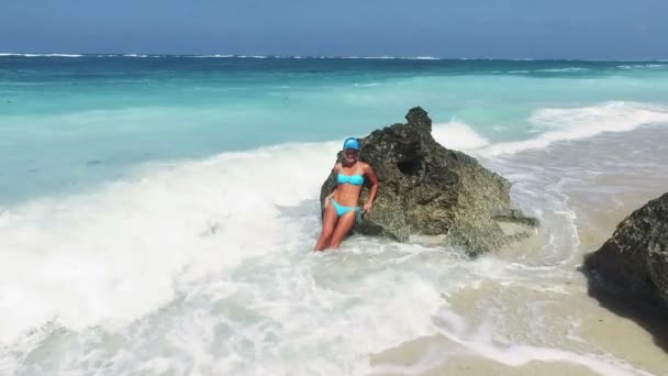 Медленное движение 100 кадров в секунду. Красивая девушка в бикини на берегу океана смеется, когда ее брызгает волна, разбивающаяся о скалы . — стоковое видео
