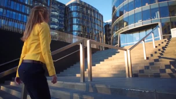 Joven chica hermosa con una chaqueta amarilla subiendo las escaleras. Mujer de negocios caminando al trabajo en el centro de negocios. Luz del sol brillante en la mañana. Vídeo de estilo cinematográfico. Clima ventoso . — Vídeo de stock