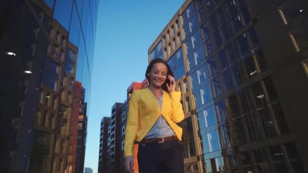 Lage hoek video bekijken van jong mooi meisje in een gele jas in de buurt van office met de telefoon. Zakenvrouw spreken door de smartphone op het business center. Fel zonlicht in de ochtend. — Stockvideo