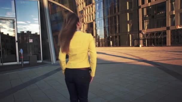 Au ralenti. Caméra suite à Jeune belle fille dans une veste jaune qui marche beatbetween grands bâtiments. Lumière du soleil vive dans la soirée. Vidéo de style cinématographique . — Video