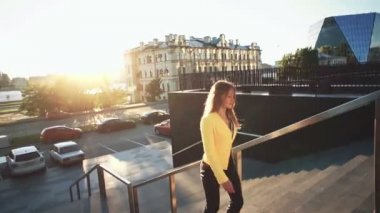 Yavaş çekim. Merdivenlerden çıkan sarı ceketli genç güzel kız. İş merkezinde işe yürüyen iş kadını. Sabahları parlak güneş ışığı. Sinematik tarzı video. Rüzgarlı hava.