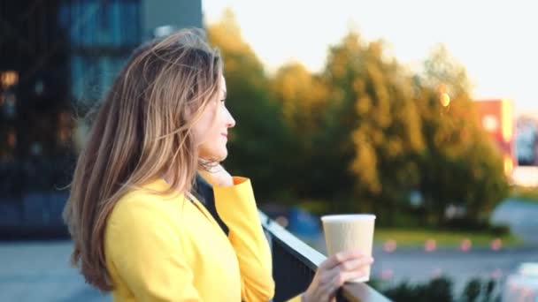 Slow motion. Ung vacker flicka i en gul jacka nära kontoret vistas och leende på balkony med kaffet. Leende affärskvinna med kopp te på affärscentret. — Stockvideo