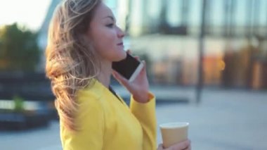 Kamera genç güzel iş kadını etrafında hareket ediyor. Esmer kız telefonla konuşuyor ve büyük şehirde ofis binası yakınında kahve içiyor. Sinematik tarzda video.