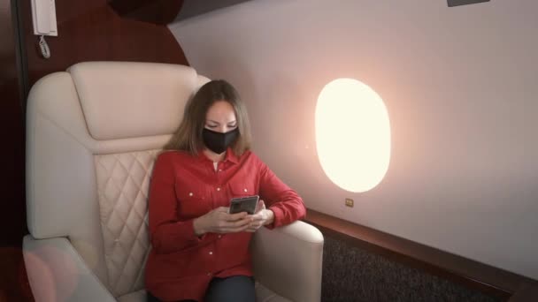 Νεαρή επιχειρηματίας φοράει μια ιατρική μάσκα κάθεται στο τζετ, κοιτάζοντας προς τα παράθυρα και χρησιμοποιώντας το κινητό τηλέφωνο. Ο ήλιος λάμπει μέσα από το παράθυρο στο αεροπλάνο. — Αρχείο Βίντεο