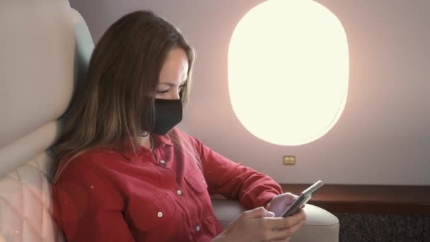 Egy fiatal üzletasszony orvosi maszkban ül a repülőn, az ablakokat bámulja és mobiltelefont használ. A gép ablakán keresztül süt a nap.. — Stock videók