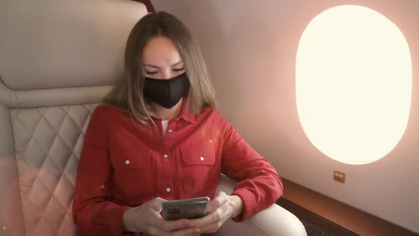 Молода ділова жінка носить медичну маску, сидячи в струмені, дивлячись у вікна і використовуючи мобільний телефон. Сонце світить крізь вікно на площині . — стокове відео