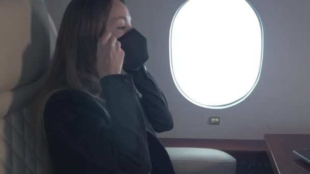 Una joven empresaria se pone una máscara médica mientras vuela en el avión de negocios. Mujer mirando a la ventana y usando smartphone. — Vídeo de stock